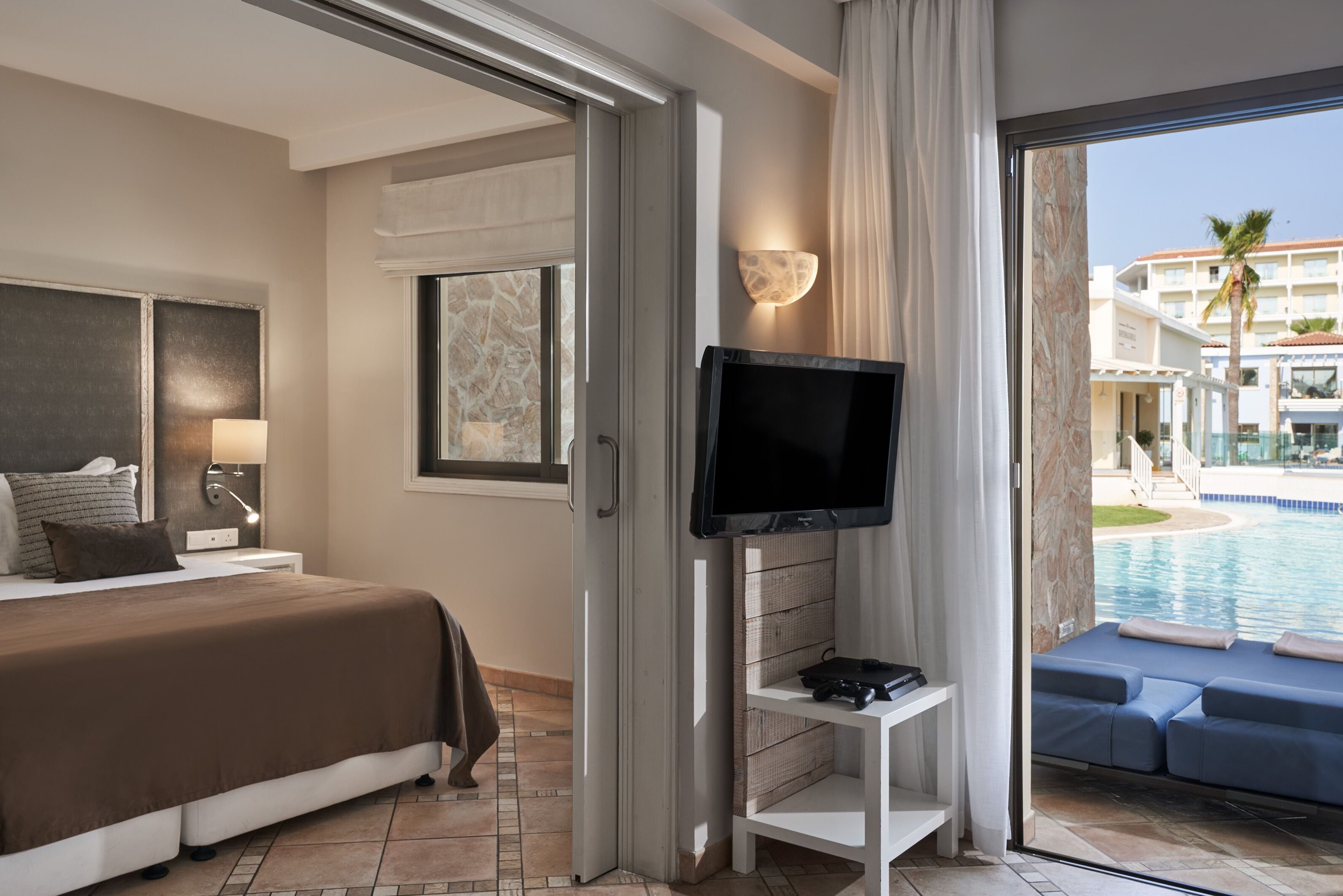 A hotel room with a bed, a TV, a small table, and a view of a pool and buildings.