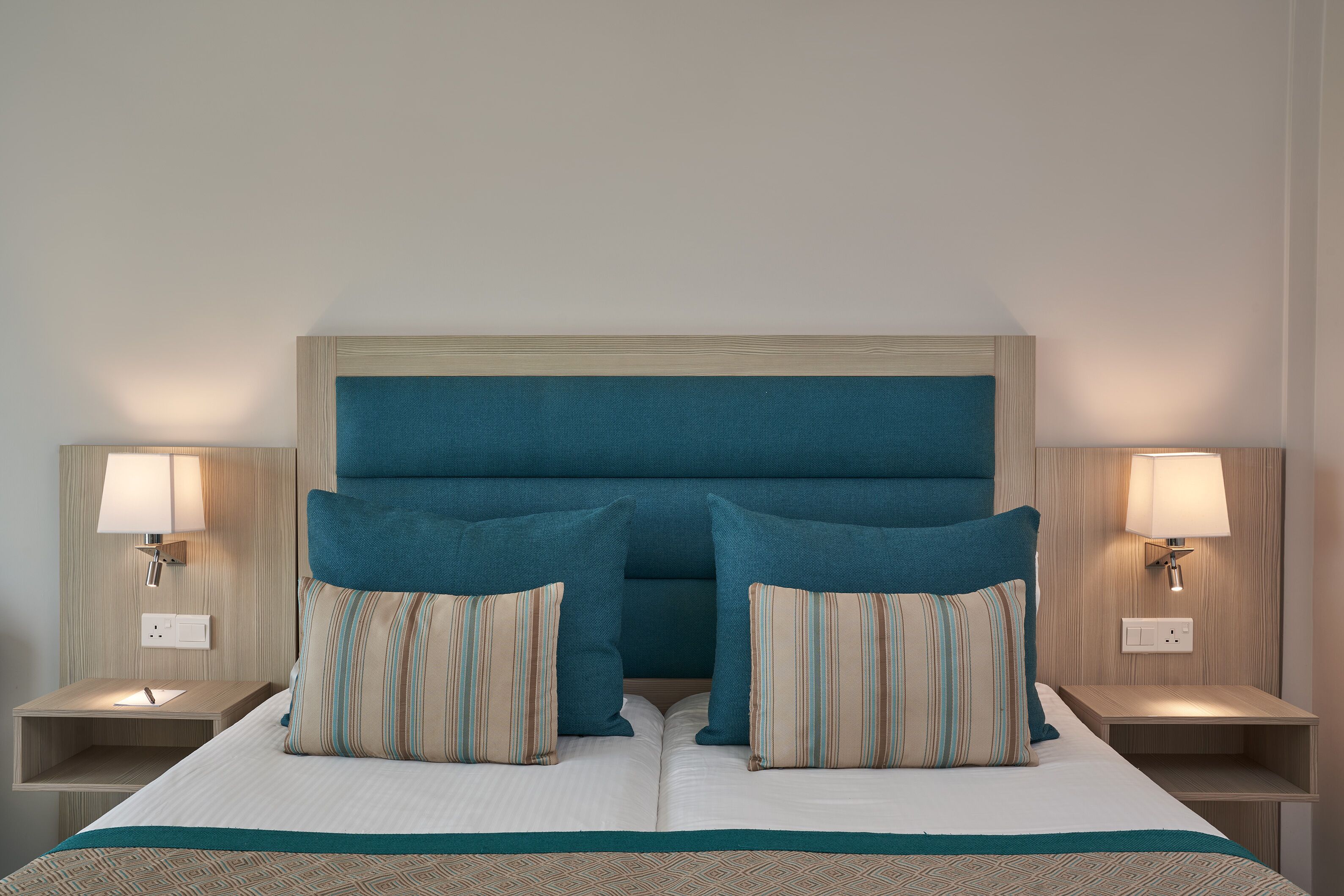 Un lit bien fait, avec un oreiller à rayures et une tête de lit de couleur turquoise. Il y a deux tables de chevet, chacune équipée d’une lampe et d’une prise électrique.