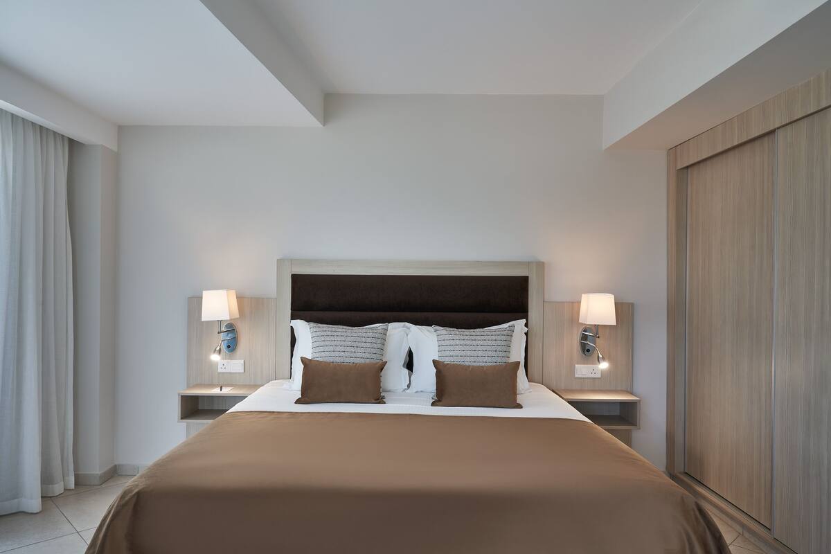 A hotel room with a large bed, two bedside tables with lamps, a wooden wardrobe, and a large window with curtains.