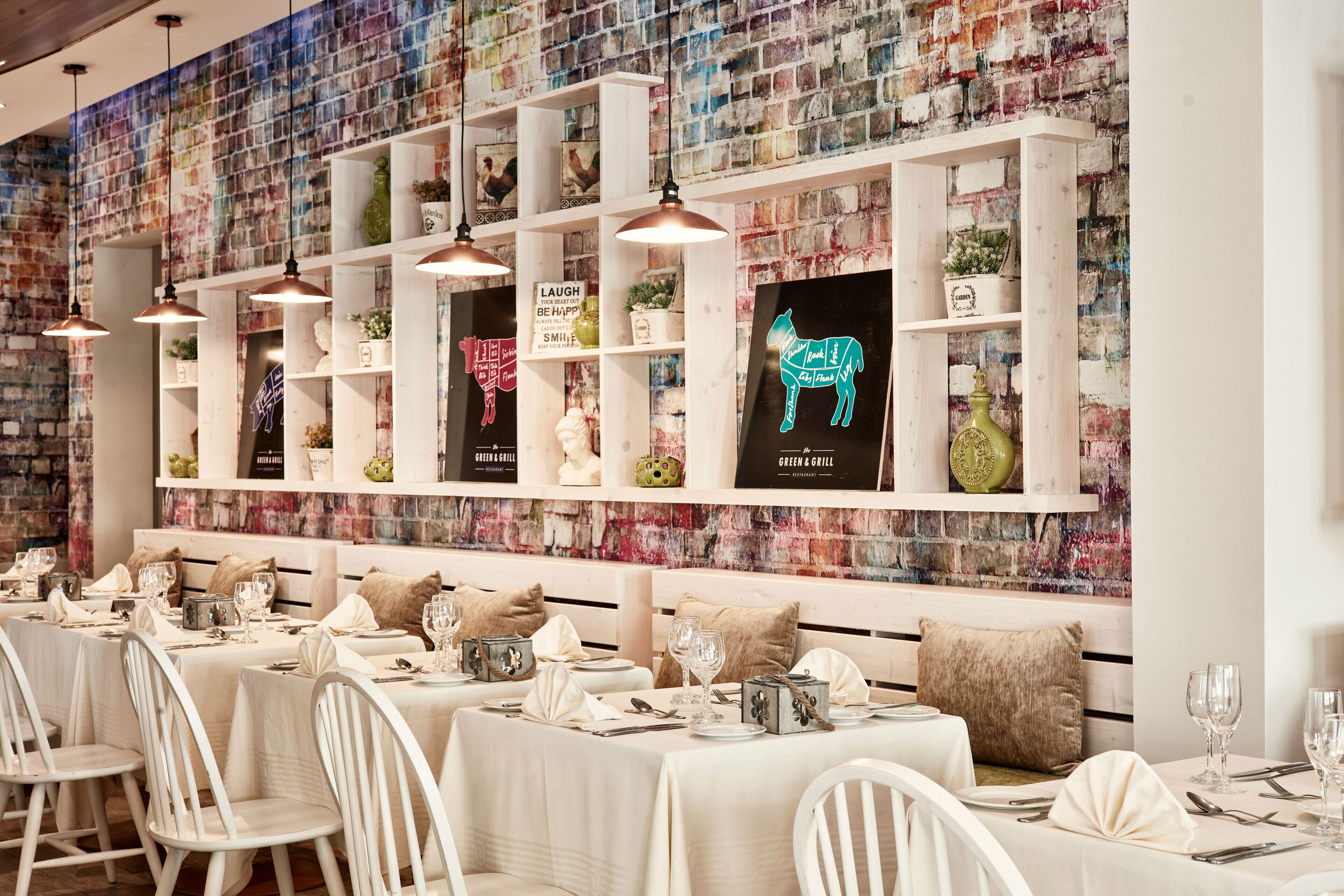 A restaurant with white tables and chairs, a brick wall with colorful artwork, and shelves with framed pictures and decorative items.