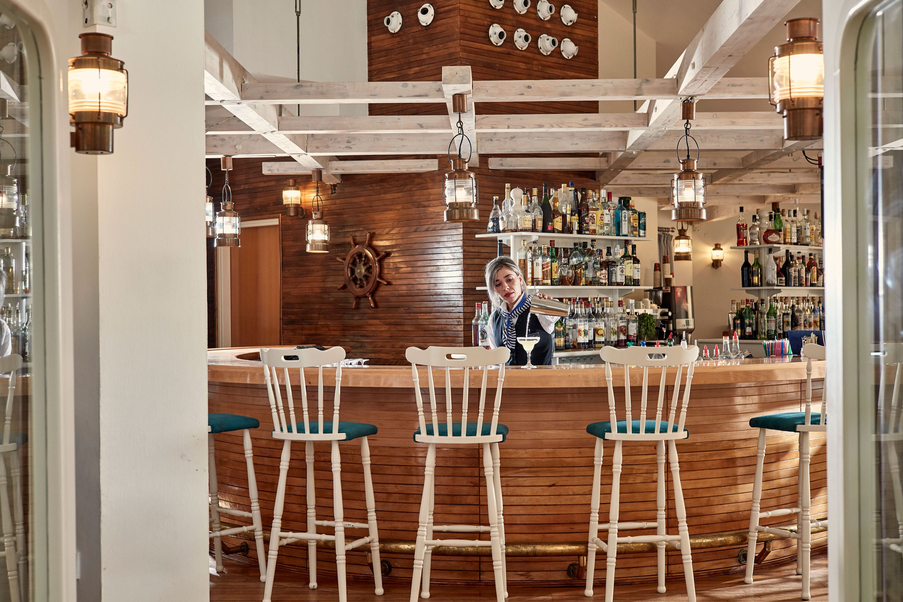 Un espace bar avec un comptoir en bois, des tabourets de bar blancs et un barman.