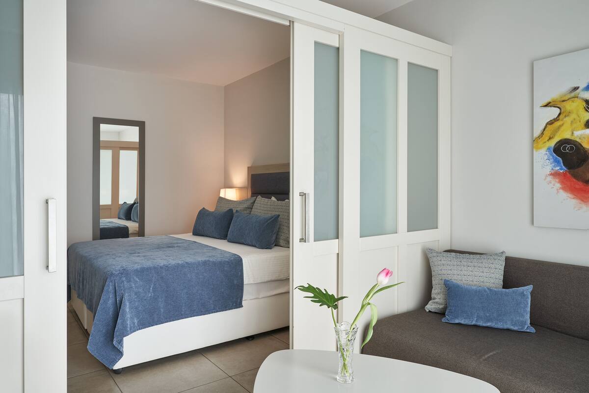 A modern bedroom with a bed, a nightstand, a vase with flowers, and a sofa with cushions.