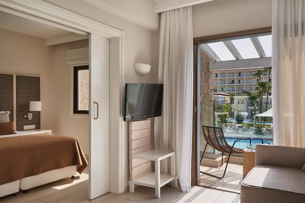 A hotel room with a bed, a TV, a small table, a chair, and a balcony with a pool view.