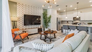 Una sala de estar moderna con un sofá, un sillón y una mesa de centro, un televisor de pantalla plana montado en la pared, y una zona de cocina con microondas y taburetes de bar.