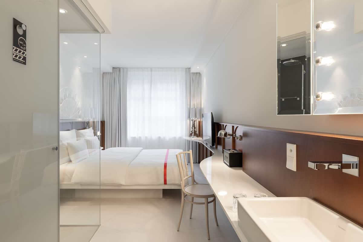 A modern hotel room with a glass partition, a bed with white linens, a wooden chair, a vanity with a mirror, and a bathtub.