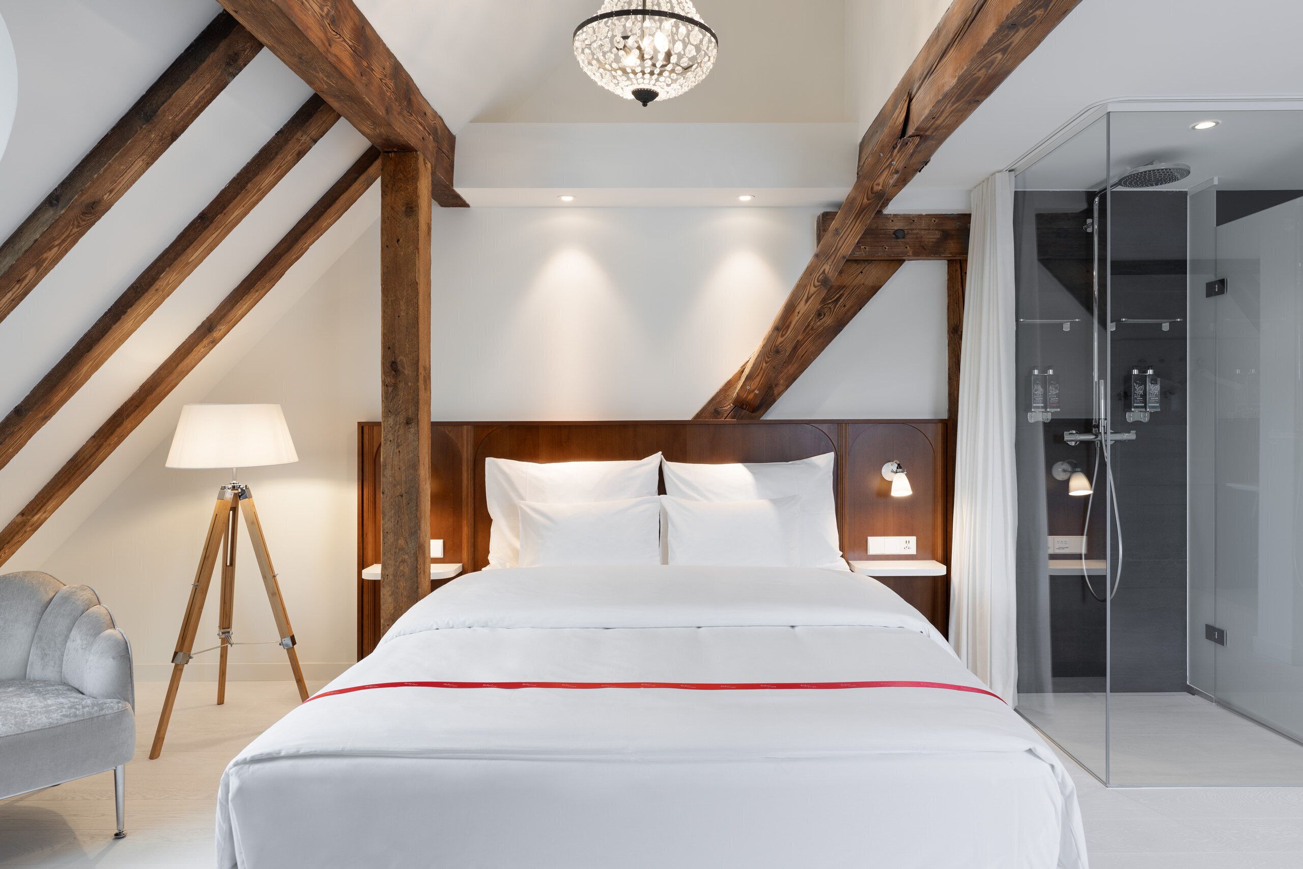 Een moderne slaapkamer met een houten plafond, een bed met witte beddengoed, een bedlamp, een vloerlamp op statief en een douchecabine met glazen wanden.