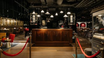 Una zona de bar con mostrador de madera, cuerdas de terciopelo rojo, lámparas colgantes y un telón rojo de fondo.
