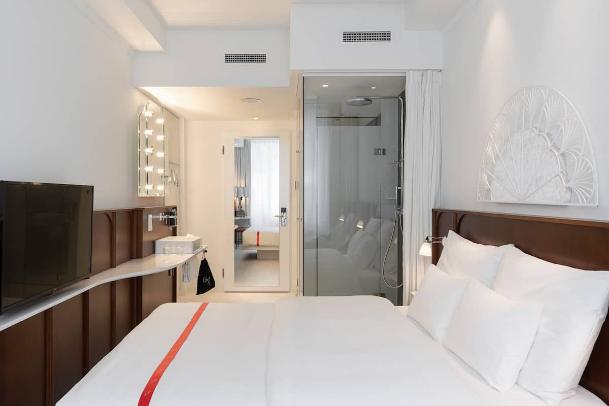 A hotel room with a bed, a TV, a mirror with lights, a shower, and a bathroom.