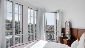 A hotel room with a large bed, a wooden nightstand, a chair, and a view of a cityscape through large windows. - Ruby Mimi Hotel Zurich by IHG (Zürich)