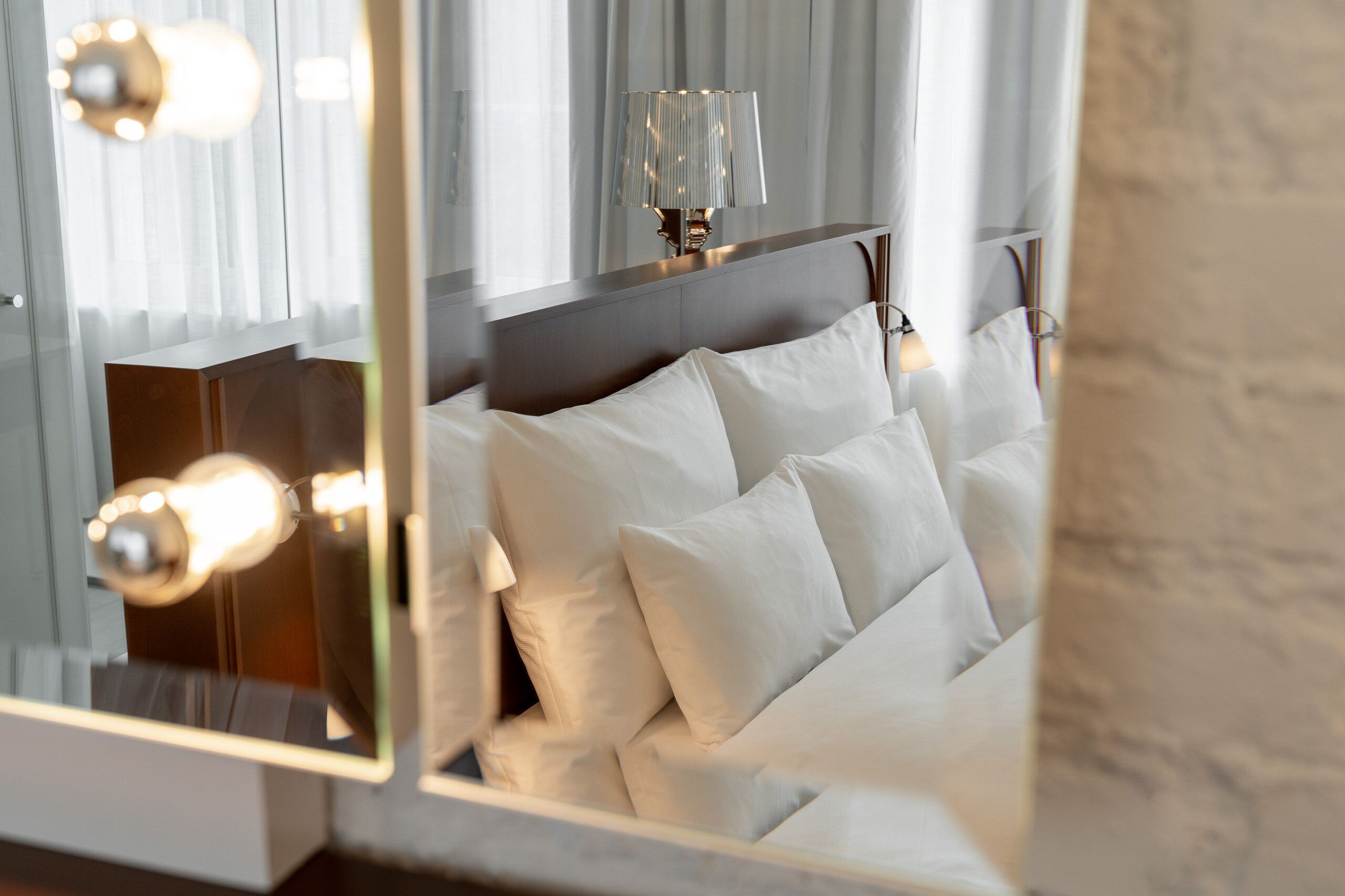 Una cama con ropa blanca, una lámpara de noche y un espejo que refleja las luminarias de la habitación.