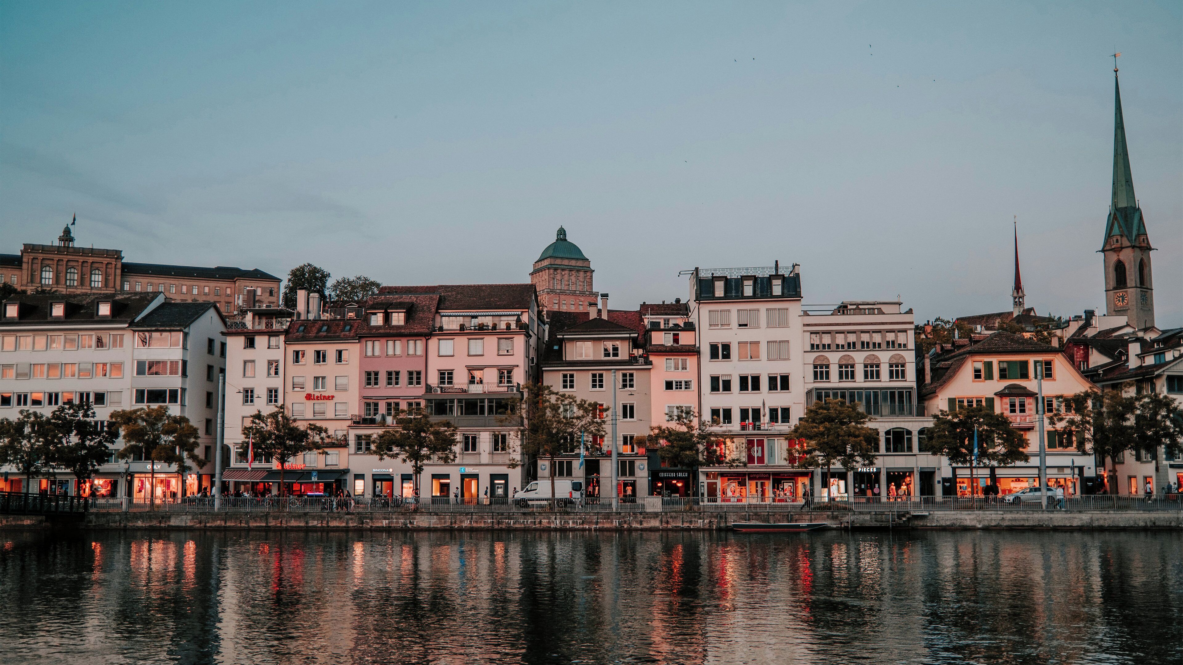 A waterfront cityscape with historic buildings, a church spire, and a river reflecting the architecture.