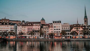 Paisagem urbana à beira-mar com edifícios históricos, uma torre de igreja e um rio refletindo a arquitetura.