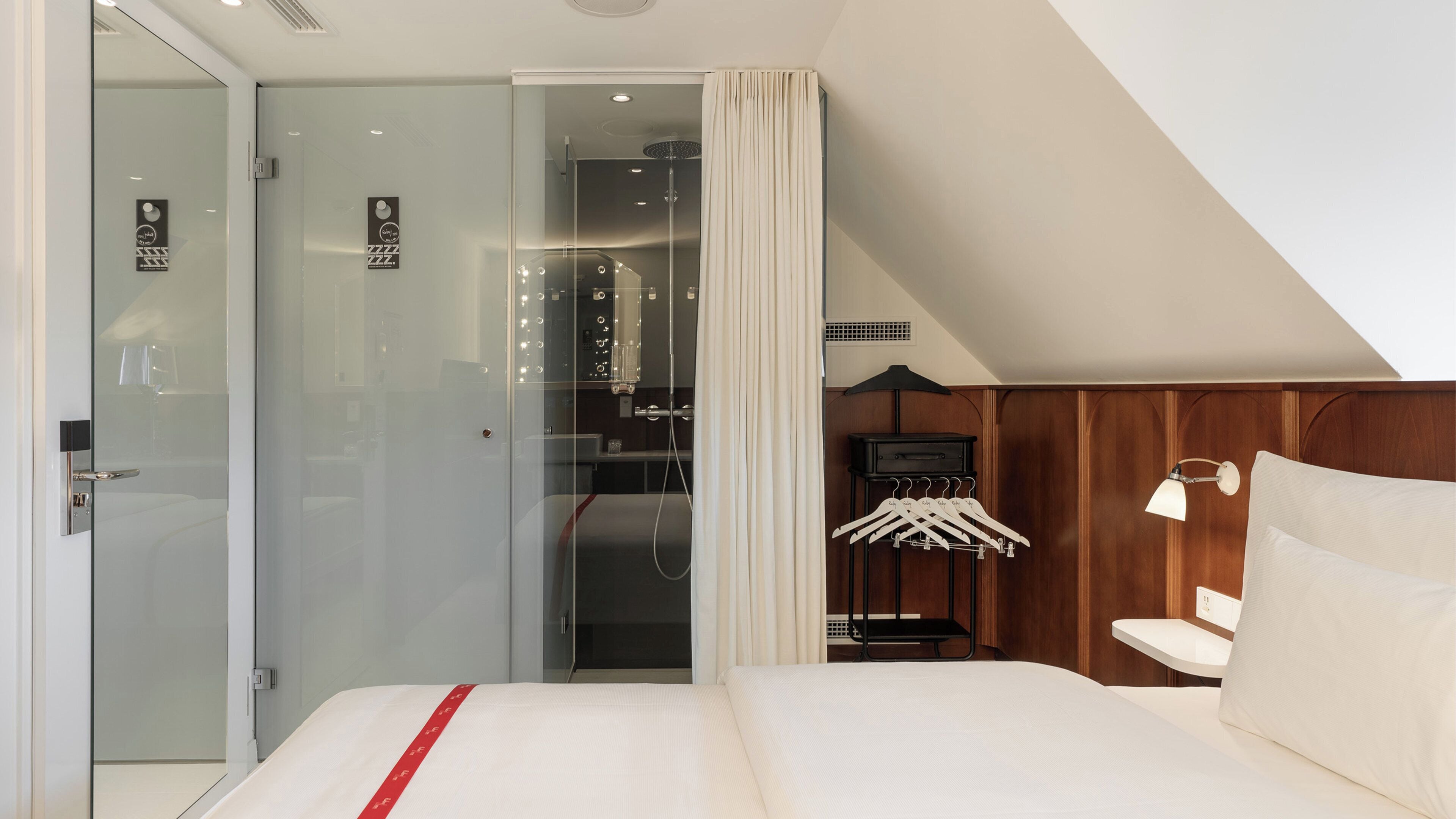 Quarto de hotel moderno com porta de vidro, cama com faixa vermelha, guarda-roupa de madeira e cabideiro.