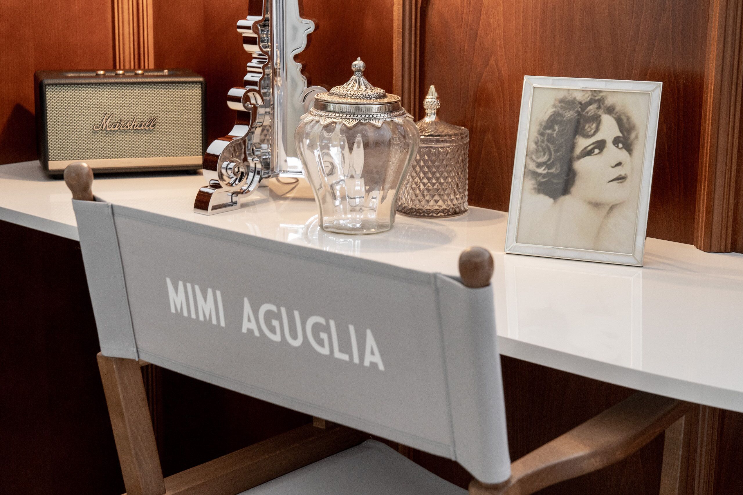 Una radio antigua, un tarro de vidrio, un cuadro enmarco y una silla con la etiqueta 'MIMI AGUGLIA' sobre una mesa blanca.