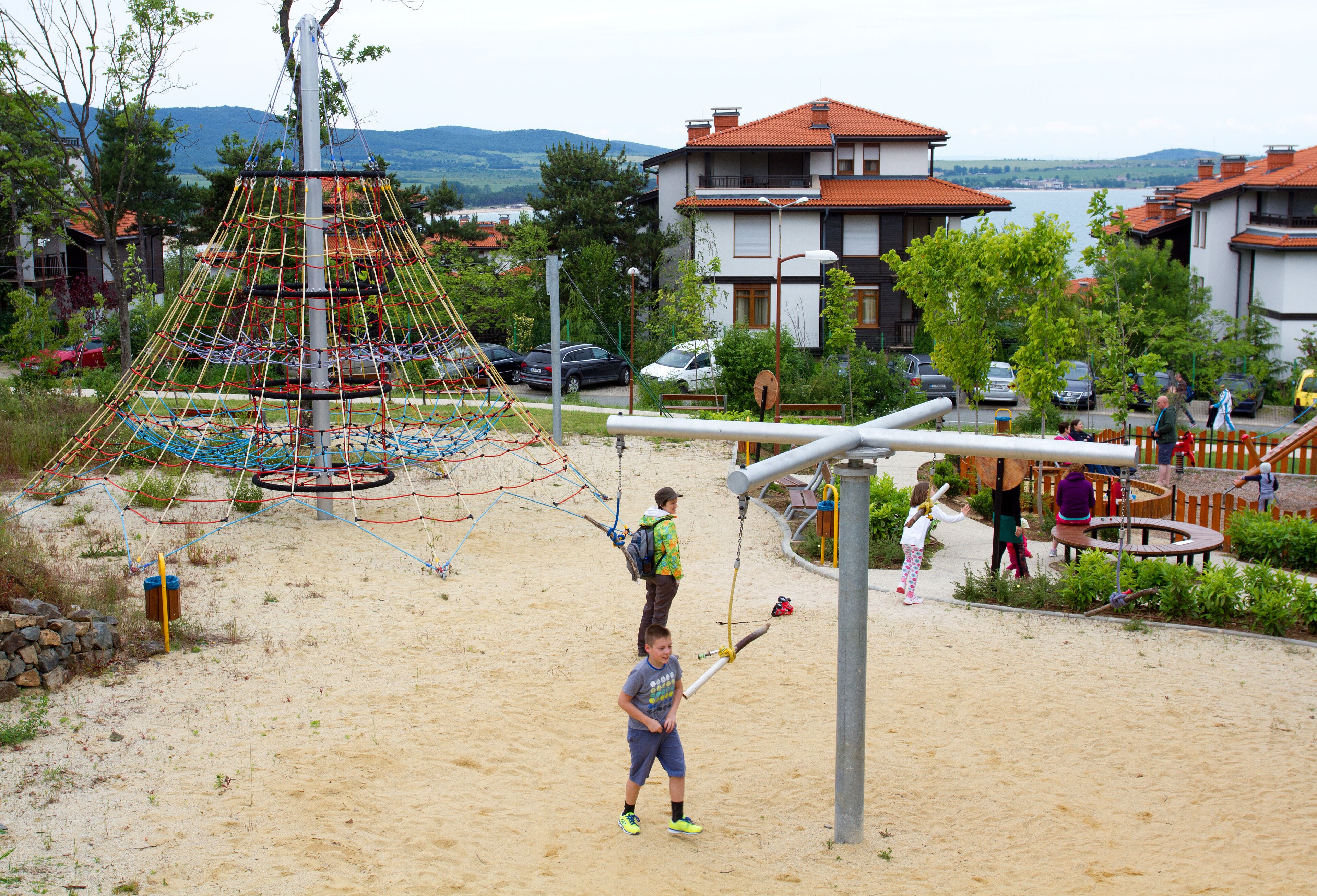 Un parque infantil con estructuras de escalada y columpios, rodeado de edificios residenciales y una masa de agua al fondo.
