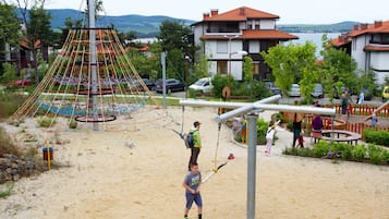 A playground with climbing structures and swings, surrounded by residential buildings and a body of water in the background.
