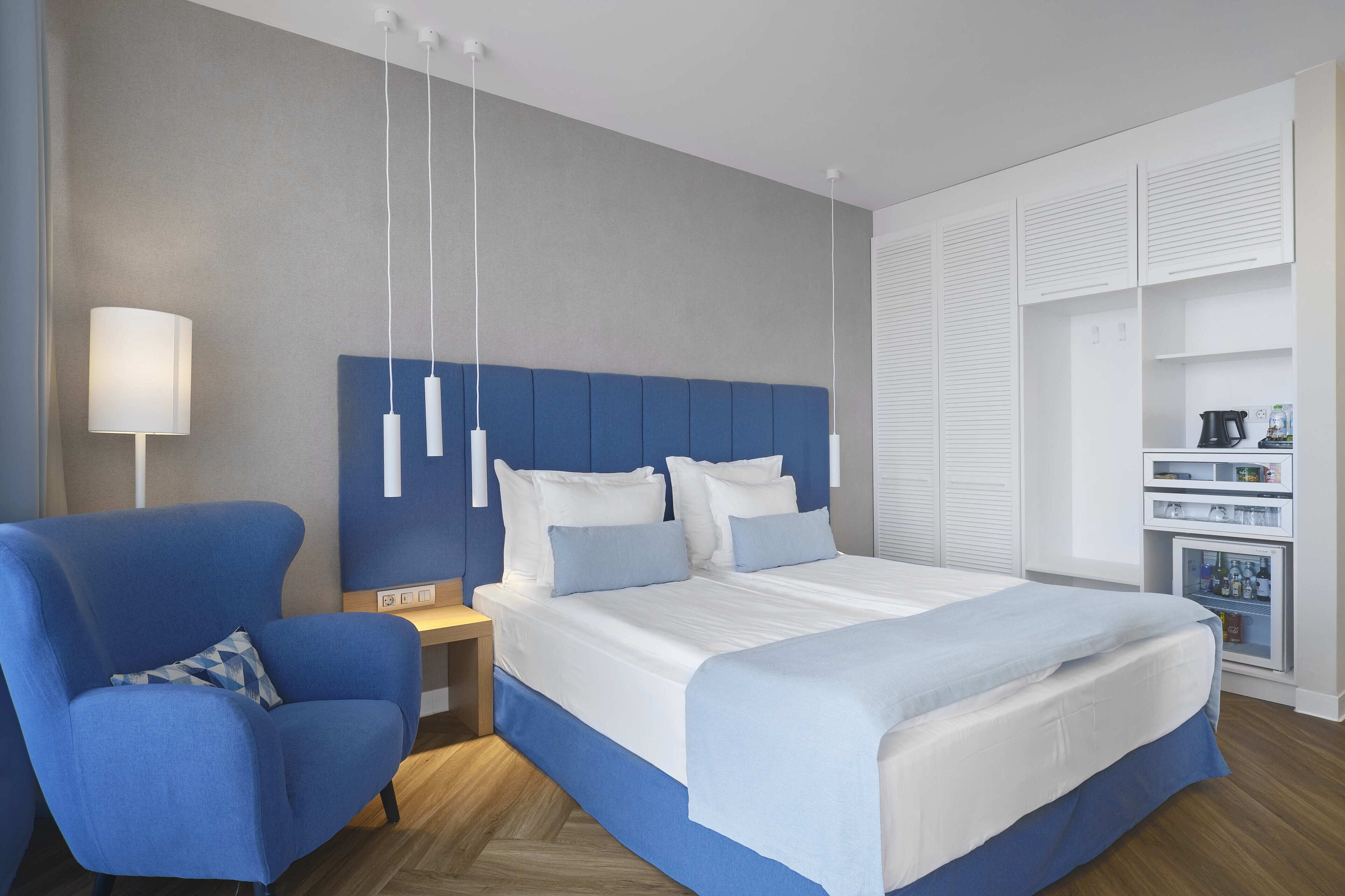 A modern hotel room with a large bed, a blue armchair, a bedside table, and a built-in wardrobe.