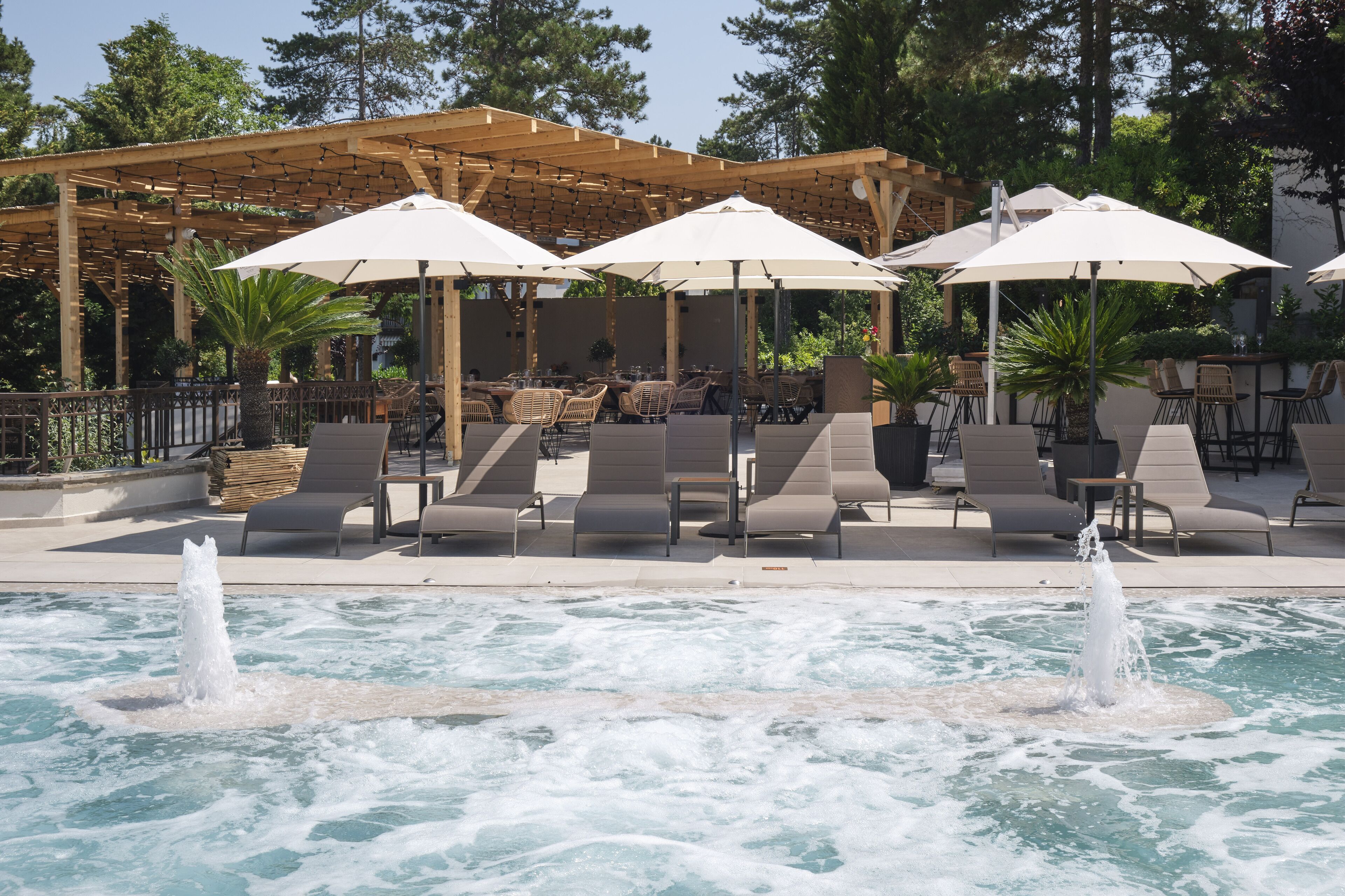 Un espace piscine équipé de chaises longues et de parasols, d’une pergola et d’une fontaine.