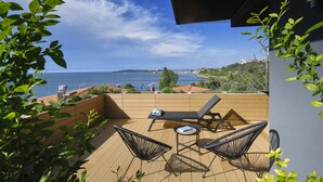 A wooden deck with two lounge chairs and a small table, overlooking a coastal view with buildings in the distance. - Viva Mare Beach Hotel (Sozopol)