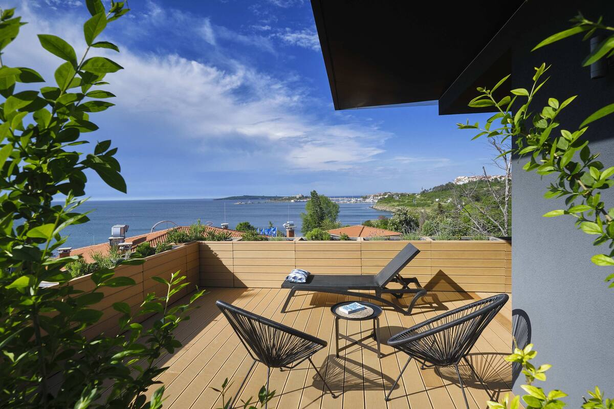 Una terraza de madera con dos sillones de descanso y una mesita, con vistas a la costa y edificios a lo lejos.