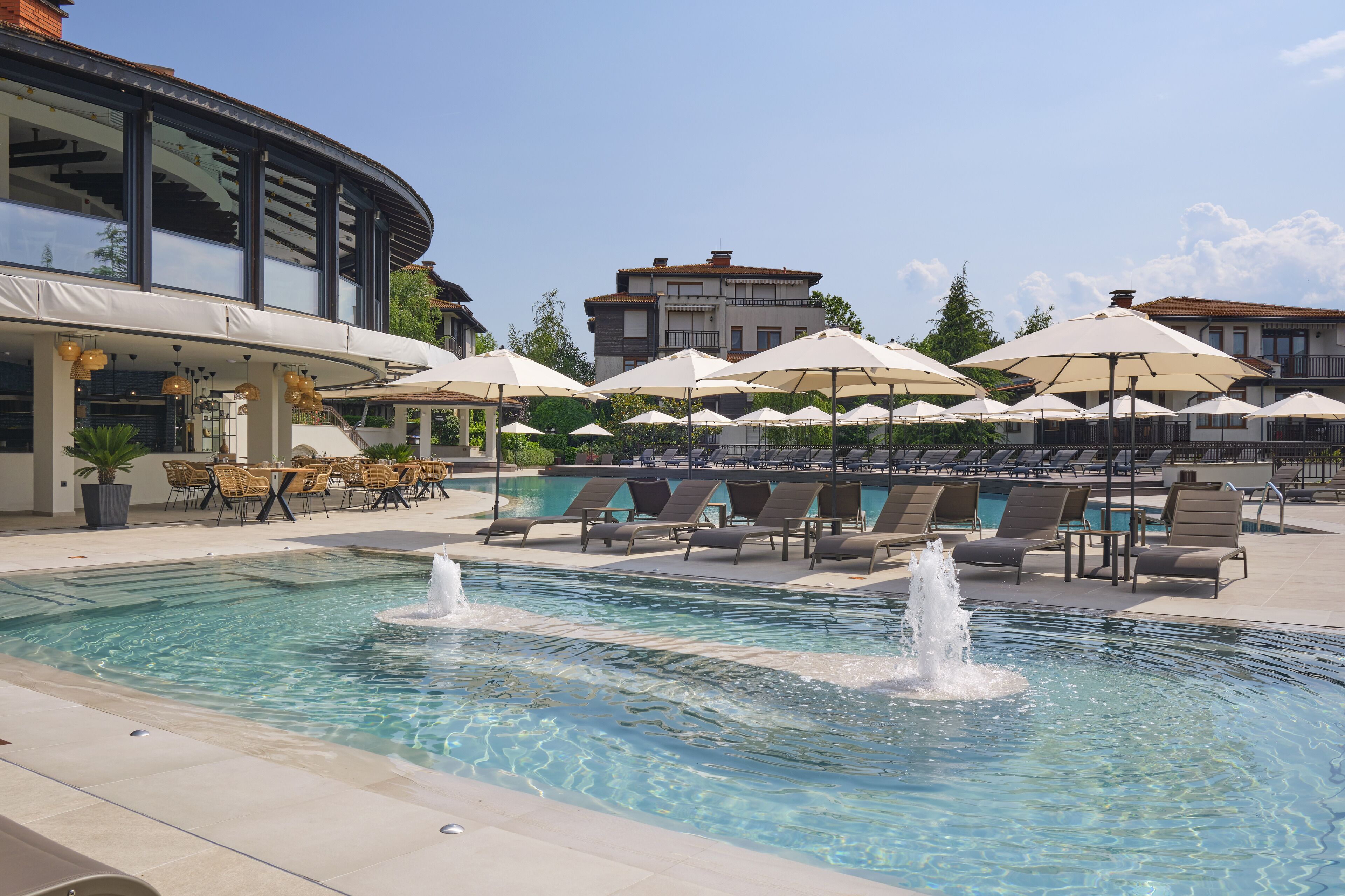 A pool area with lounge chairs and umbrellas, a fountain, and a building with large windows in the background.