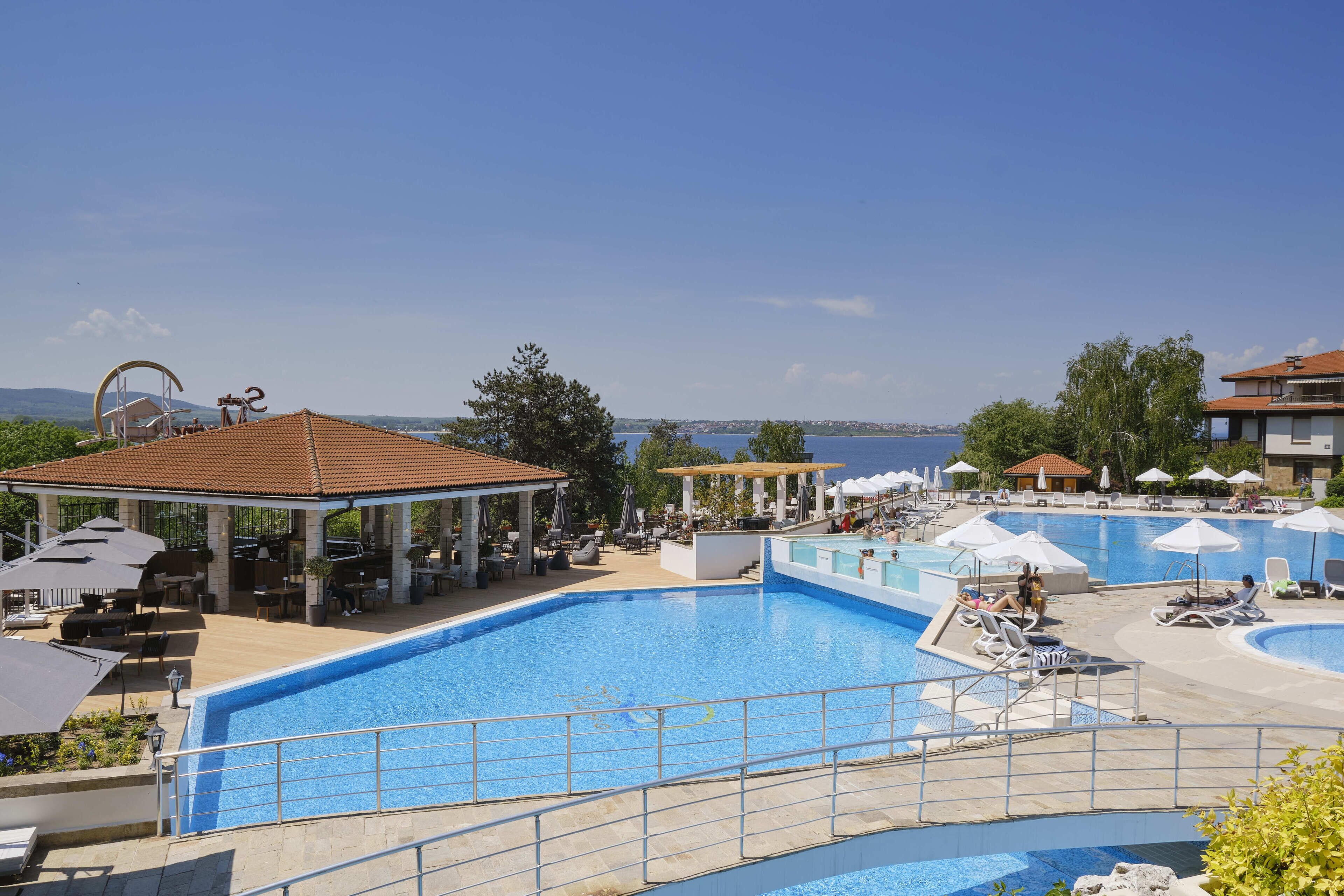 A large outdoor swimming pool with lounge chairs and umbrellas, surrounded by buildings and a view of the sea.