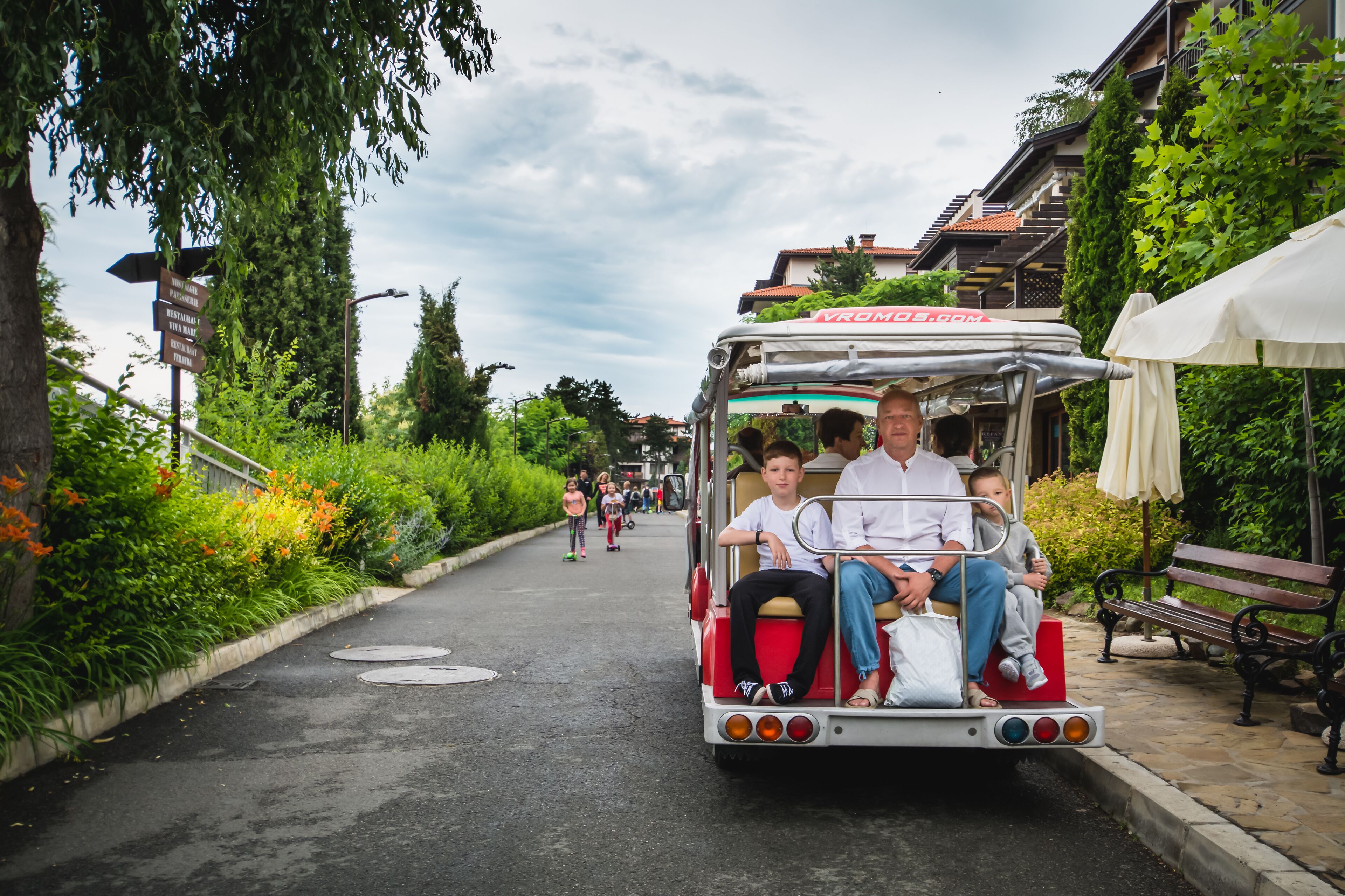 A vintage trolley with passengers, parked on a cobblestone path lined with greenery and benches.
