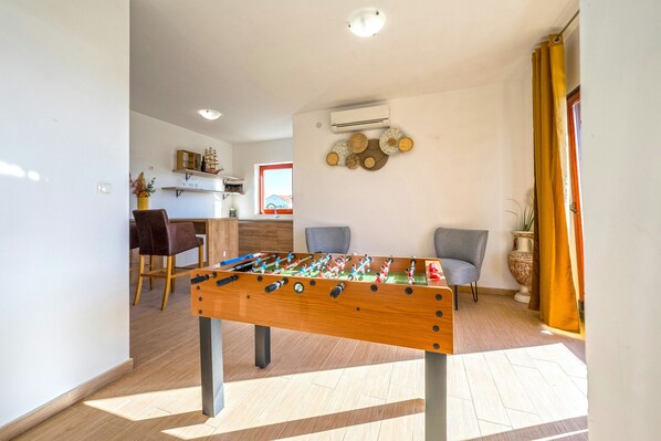 A room with a foosball table, a dining area with chairs, and a kitchen in the background.