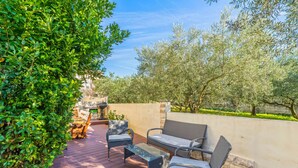 An outdoor patio with wicker furniture, a stone wall, and olive trees in the background.