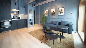 A modern kitchen with dark cabinetry, a dining area with a round table and chairs, and a living room with a sofa and a small side table. - Home Again Kirkegata 30 (Stavanger)
