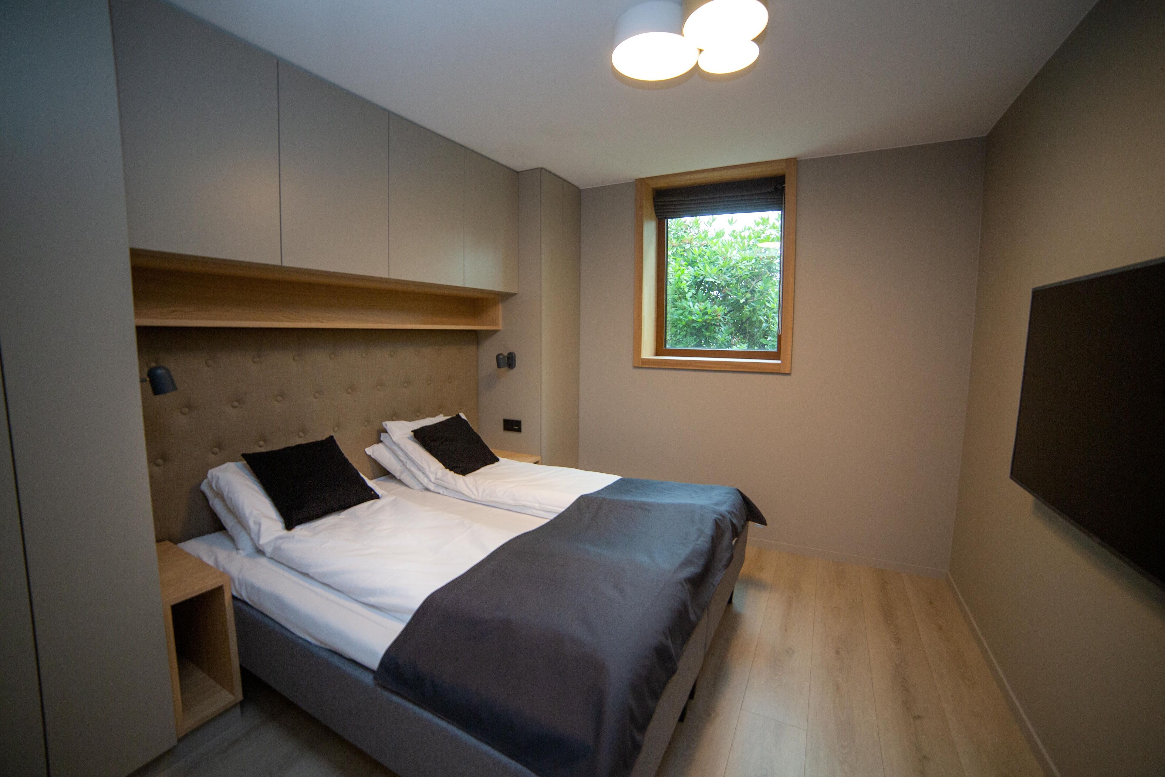 Een slaapkamer met een bed, een vastgezet hoofdbord, nachtkastjes en een raam met uitzicht op groen.