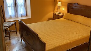A bedroom with a wooden bed, a nightstand with a lamp, a chair, a window with curtains, and a painting on the wall. - Studio (st crepin carlucet)