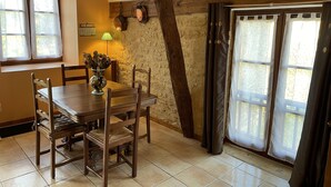 A dining area with a wooden table, chairs, and a vase of dried flowers. There is a lamp on the wall and a window with curtains. - Studio (st crepin carlucet)