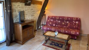 A cozy living room with a stone wall, wooden beams, a TV on a wooden cabinet, a patterned sofa, and a small coffee table. - Studio (st crepin carlucet)
