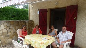 Eine Gruppe von Personen sitzt an einem runden Tisch mit gelber Tischdecke, umgeben von weißen Plastikstühlen, auf einer Terrasse mit Steinmauern und roten Fensterläden.