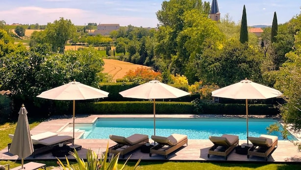 A pool area with lounge chairs and umbrellas, surrounded by greenery and a distant church.