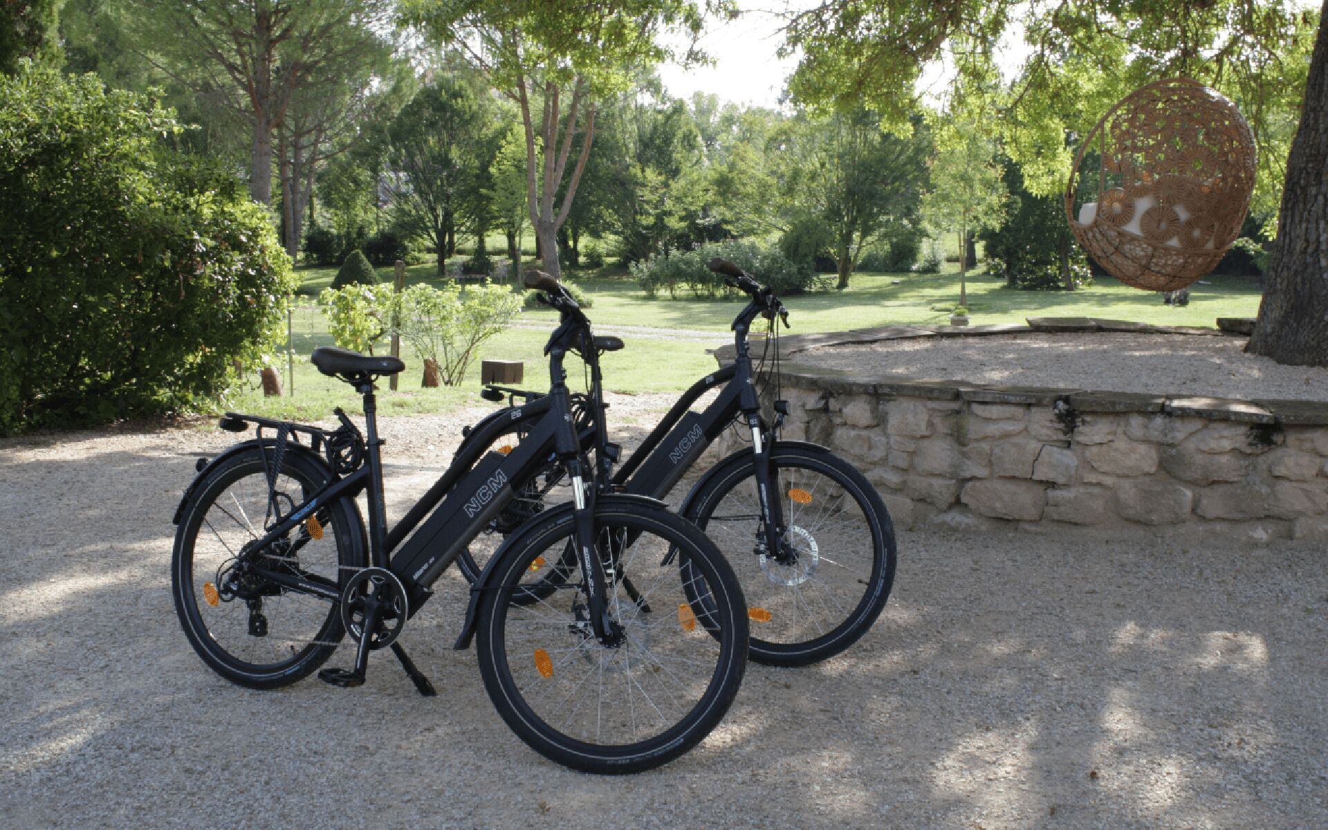 Deux vélos sont garés sur un chemin de gravier dans un parc, avec des arbres et un mur de pierre en arrière-plan.