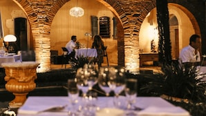An outdoor dining area with brick arches, tables set with white tablecloths and wine glasses, and a chandelier hanging above.