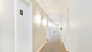 Un lungo e stretto corridoio d'albergo con pareti bianche, pavimento rivestito di moquette e una serie di porte su un lato.
