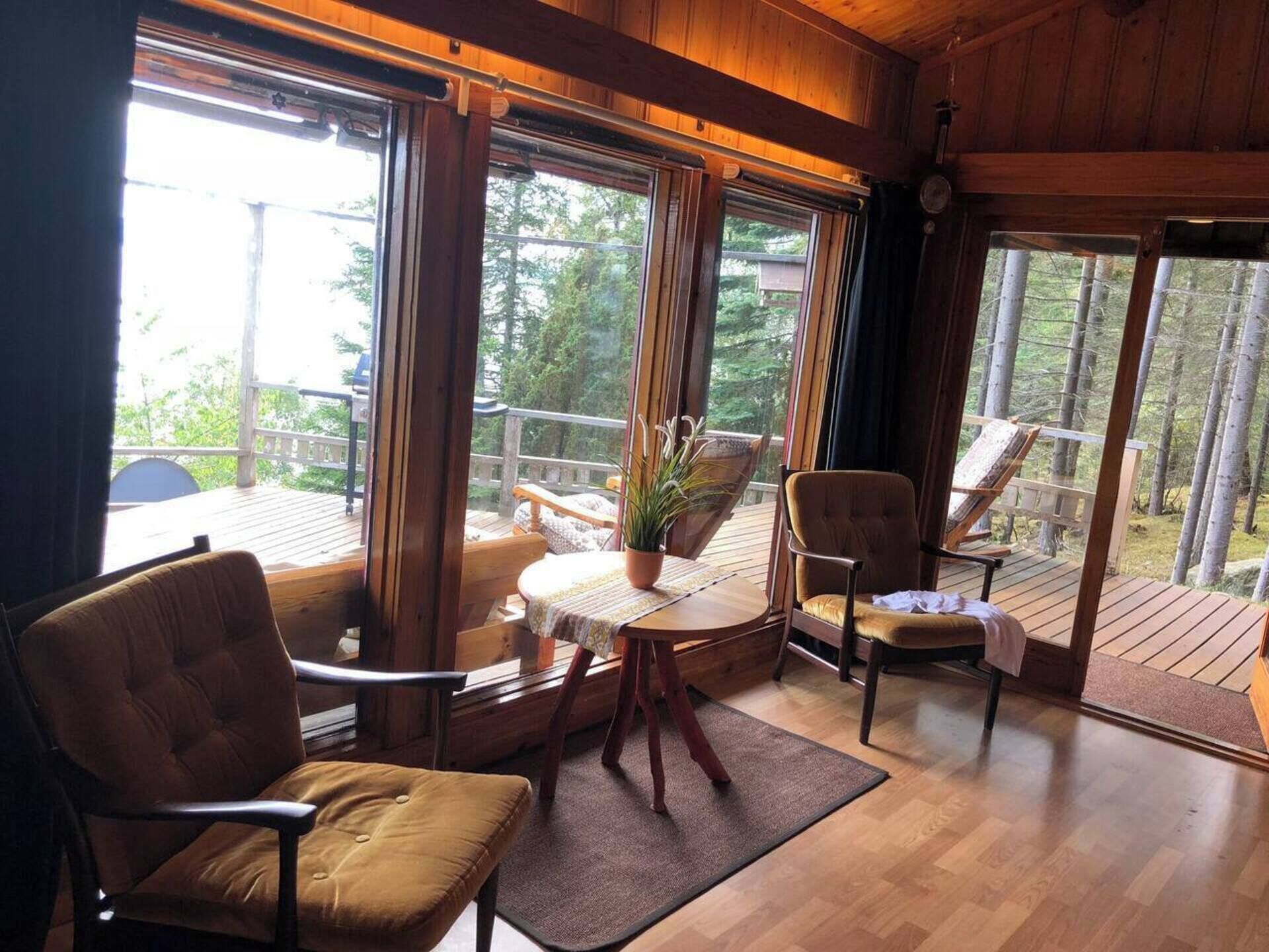 A cozy room with a wooden floor, a table with chairs, a potted plant, and large windows offering a view of trees.