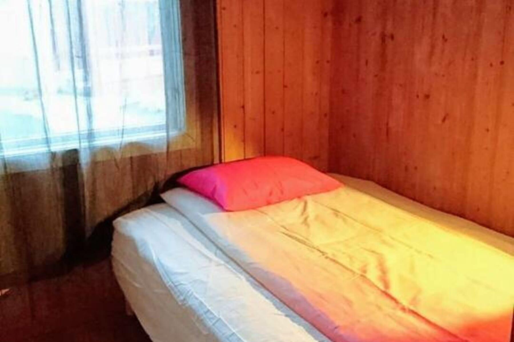 A single bed with a pink pillow and white bedding in a room with wooden walls and a window with sheer curtains.
