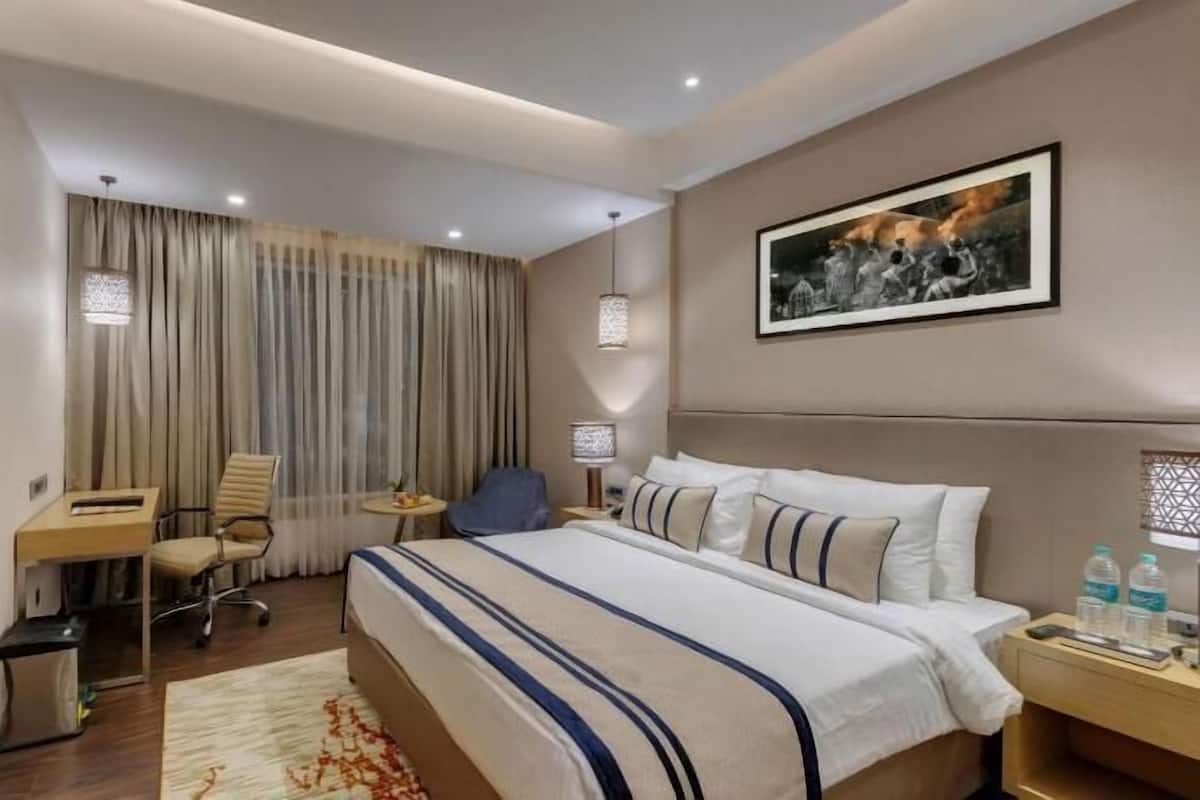 A hotel room with a bed, a desk, a chair, a small table, a TV, and a painting on the wall.