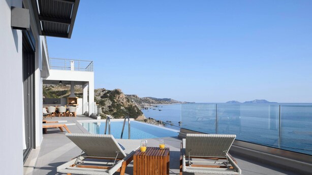 A modern outdoor terrace with two wicker lounge chairs, a wooden table, and a clear view of the sea and rocky coastline. - Rozites Luxury Beachfront Villa, By Hellocrete (Agia Foteini)