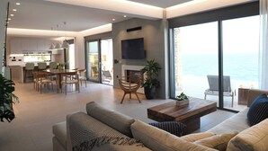 A modern living room with a large sofa, a coffee table, and a dining area with chairs and a table. There is a large window with a view of the ocean. - Rozites Luxury Beachfront Villa, By Hellocrete (Agia Foteini)