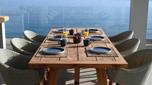 A wooden dining table with blue plates, glasses, and a bowl of fruit, set on a balcony with wicker chairs and a view of the sea. - Rozites Luxury Beachfront Villa, By Hellocrete (Agia Foteini)