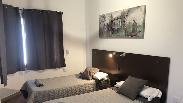A hotel room with two beds, a large window with curtains, a painting on the wall, and a headboard with lights. - TAMONANTE HOSTEL (Gran Tarajal)