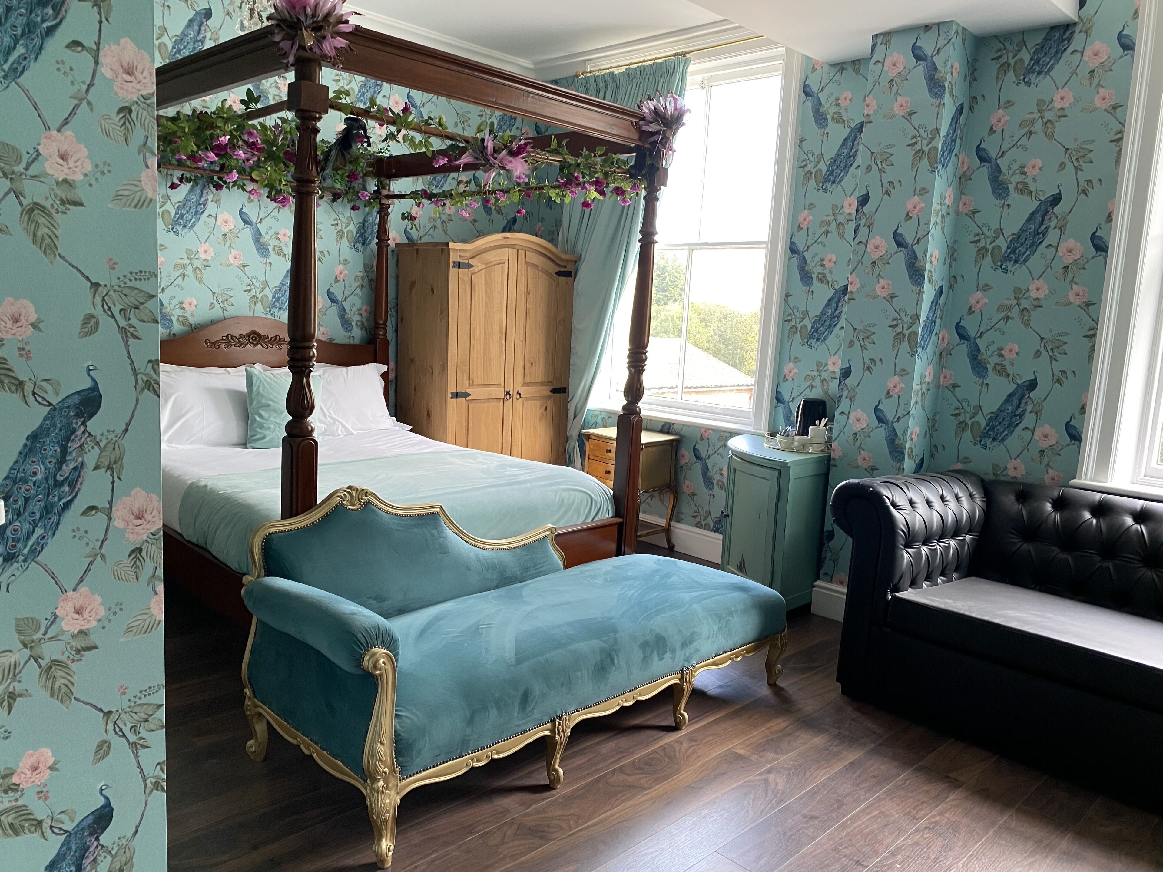 A bedroom with a four-poster bed, a vanity, a black leather sofa, and floral wallpaper.