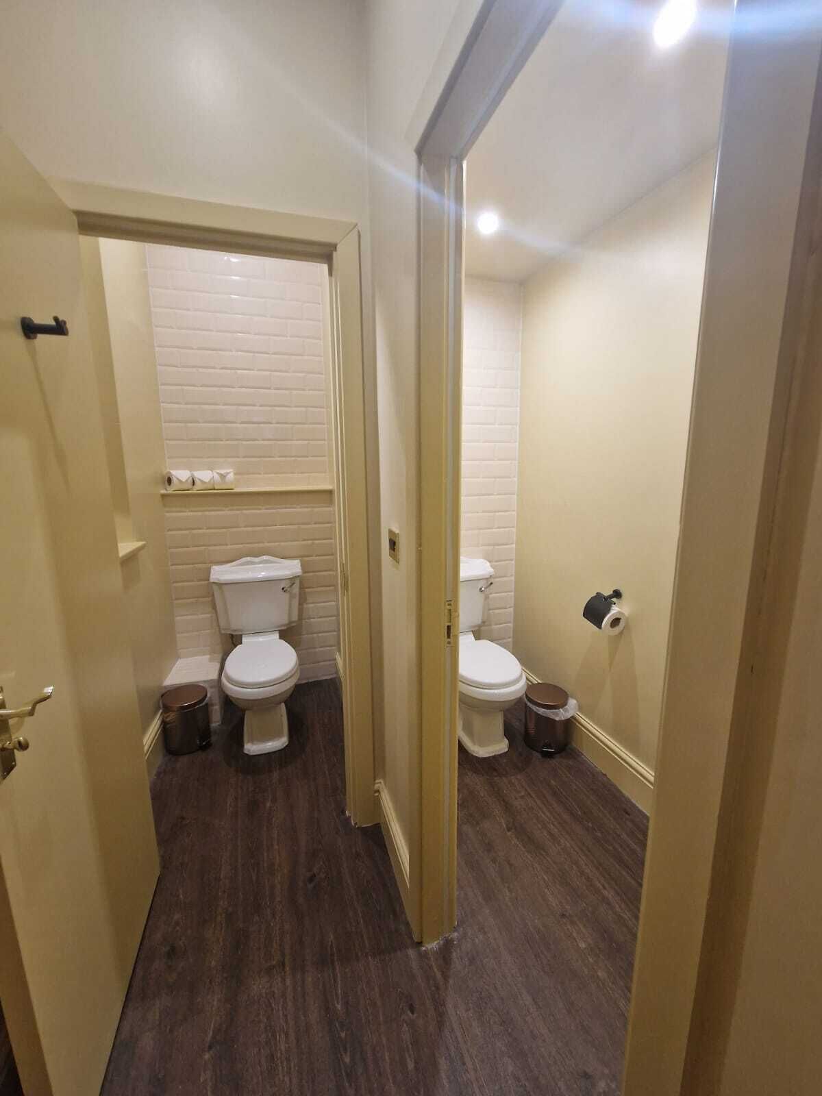 Un baño pequeño y funcional con dos inodoros, un lavamanos y un cubo de basura.