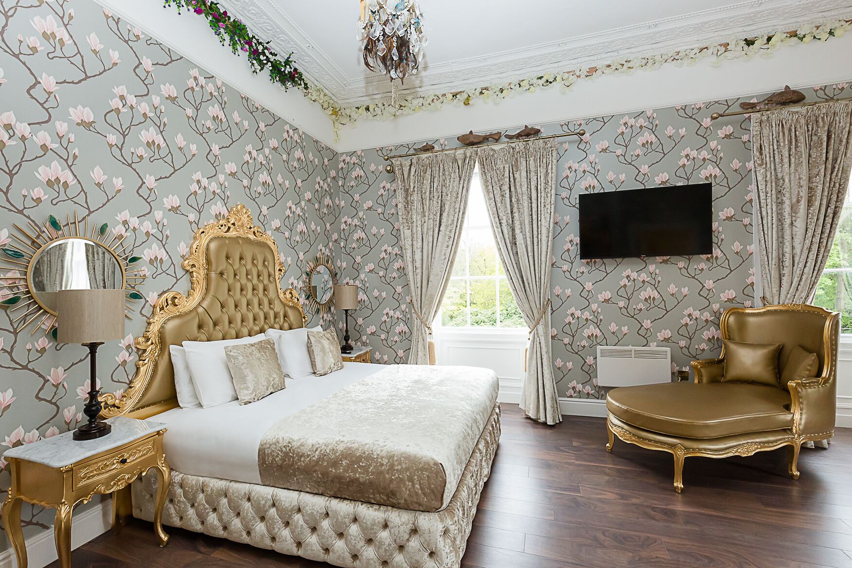 A luxurious bedroom with a large bed, a gold-framed headboard, a gold armchair, and a television on the wall.