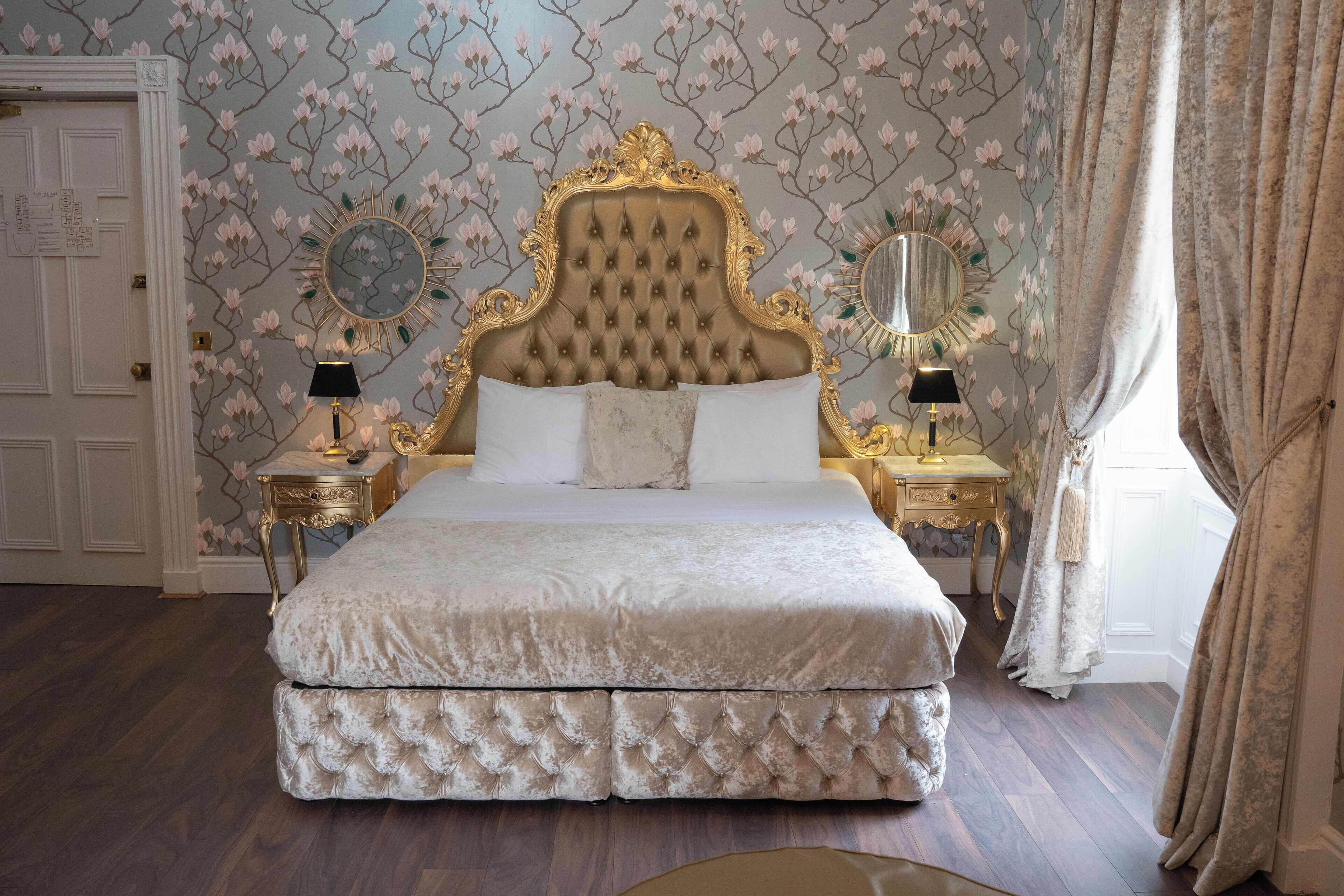 Ein großes, verziertes Bett mit einer gepolsterten Kopfteil, begleitet von zwei Nachttischen mit Lampen und Spiegeln, vor einem floralen Wandhintergrund.